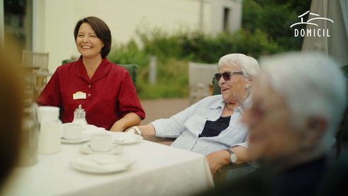 Sandra Ruppert sitzt lachend im Garten Ihrer Einrichtung zusammen mit Bewohnern an einem Kaffeetisch.