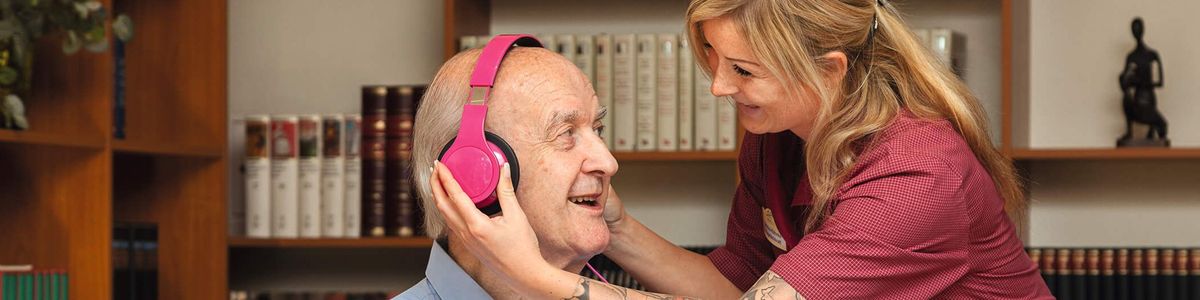 Eine Altenpflegerin setzt einem Bewohner einer DOMICIL Seniorenresidenz einen Kopfhörer auf. Der ältere Herr sitzt in einer Bibliothek und hält als Abspielgerät ein Mobiltelefon in der Hand.  