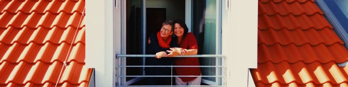 Wohnbereichsleiterin Sandra Ruppert steht mit einer Bewohnerin freundschaftlich umarmt an einem geöffneten Fenster.